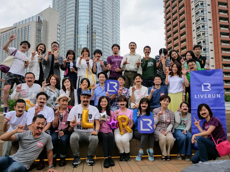 A large LIVERUN member gathering on a Tokyo rooftop, with a LIVERUN banner and LR letter props