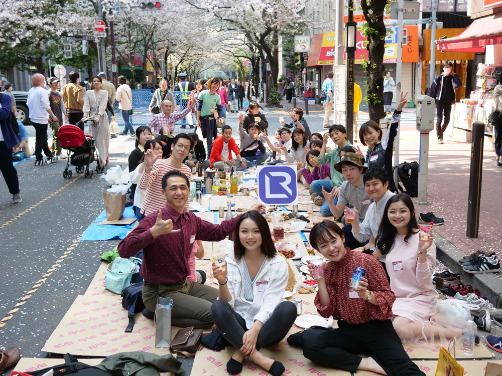 LIVERUN members at a member-organised hanami picnic on a closed Tokyo street, seated on mats beneath cherry blossom with a LIVERUN board held up